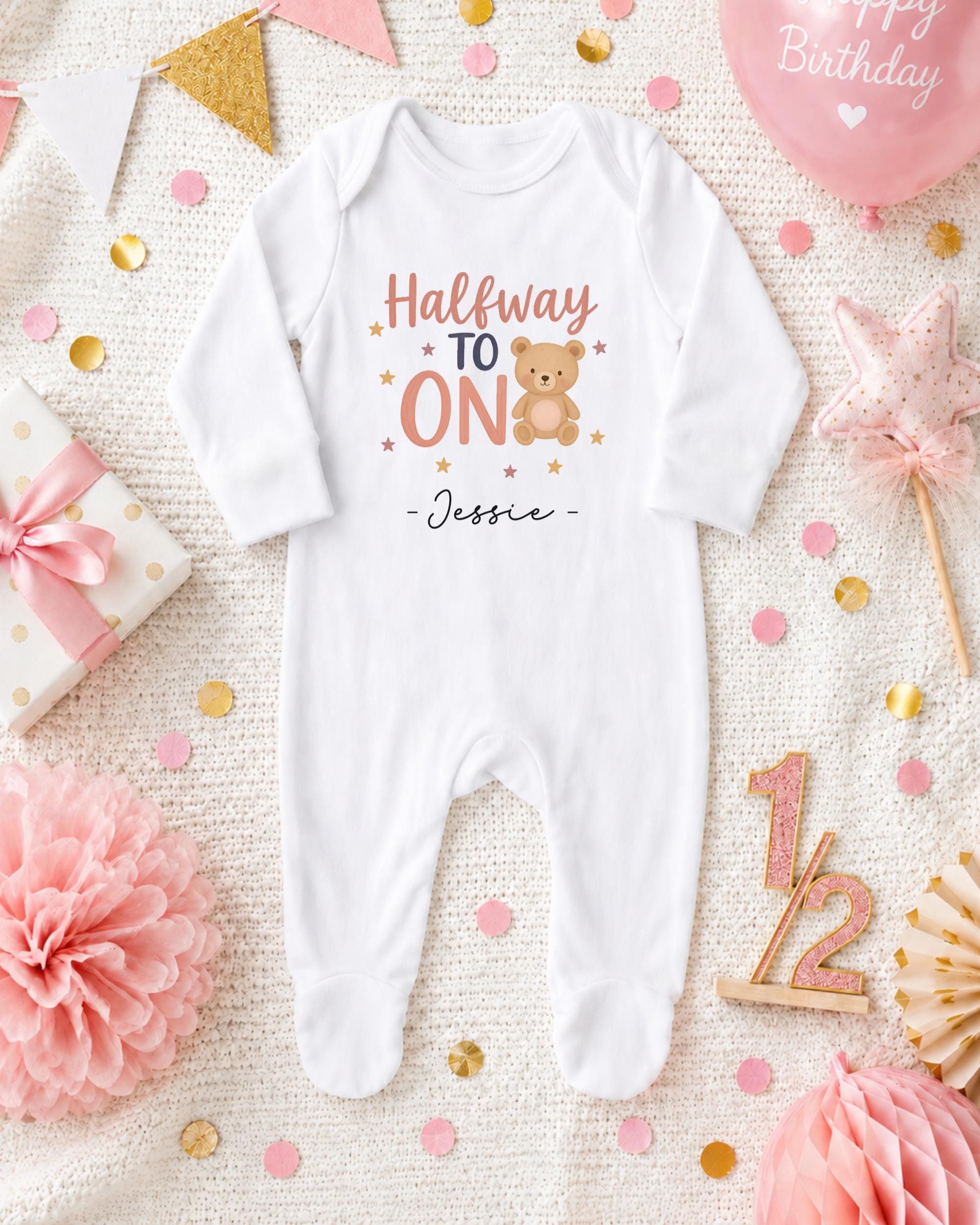 A white onesie with the text "Halfway to one" and a teddy bear graphic, surrounded by confetti, a pink balloon, and other baby-related items.