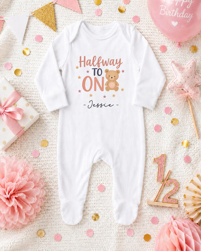 A white onesie with the text "Halfway to one" and a teddy bear graphic, surrounded by confetti, a pink balloon, and other baby-related items.