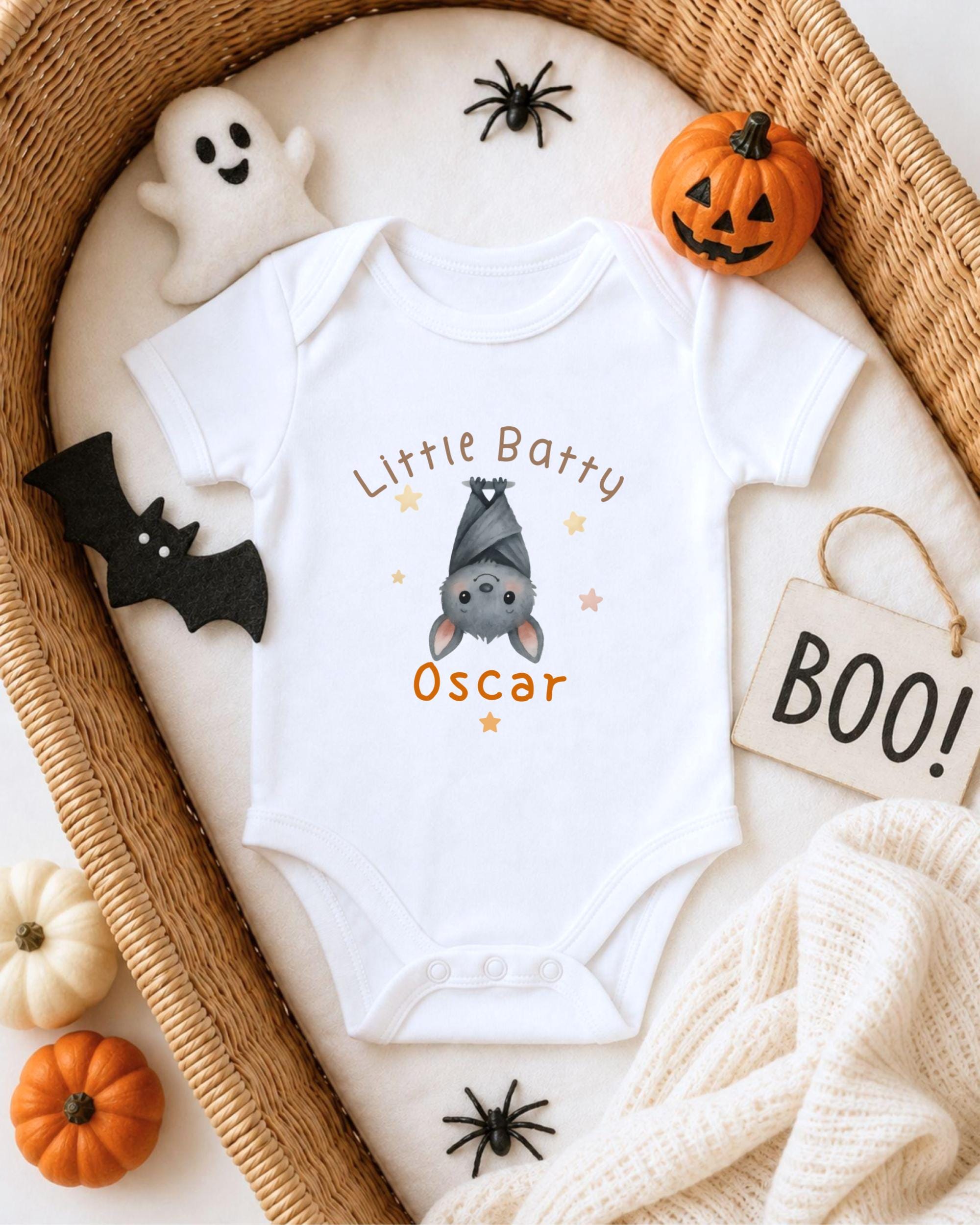 A white onesie with a bat design and the name "Oscar" printed on it, surrounded by Halloween-themed items such as a ghost, a pumpkin, a bat, and a sign that says "BOO!".
