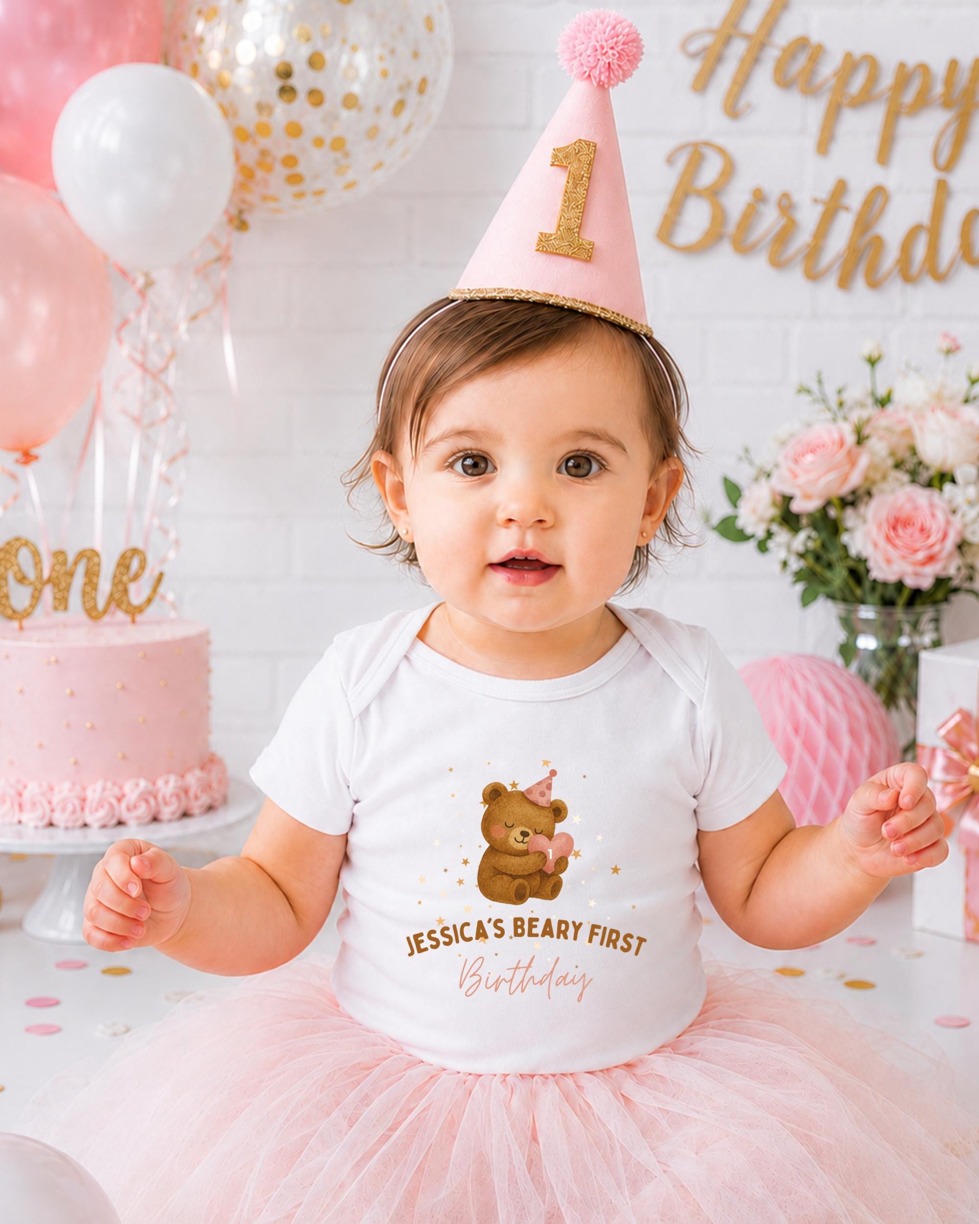 A young girl wearing a birthday hat and a white t-shirt with a teddy bear design is posing in front of a birthday cake and balloons.