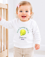 A smiling baby girl wearing a white sweatshirt with a tennis ball and text, standing next to a wooden crib.