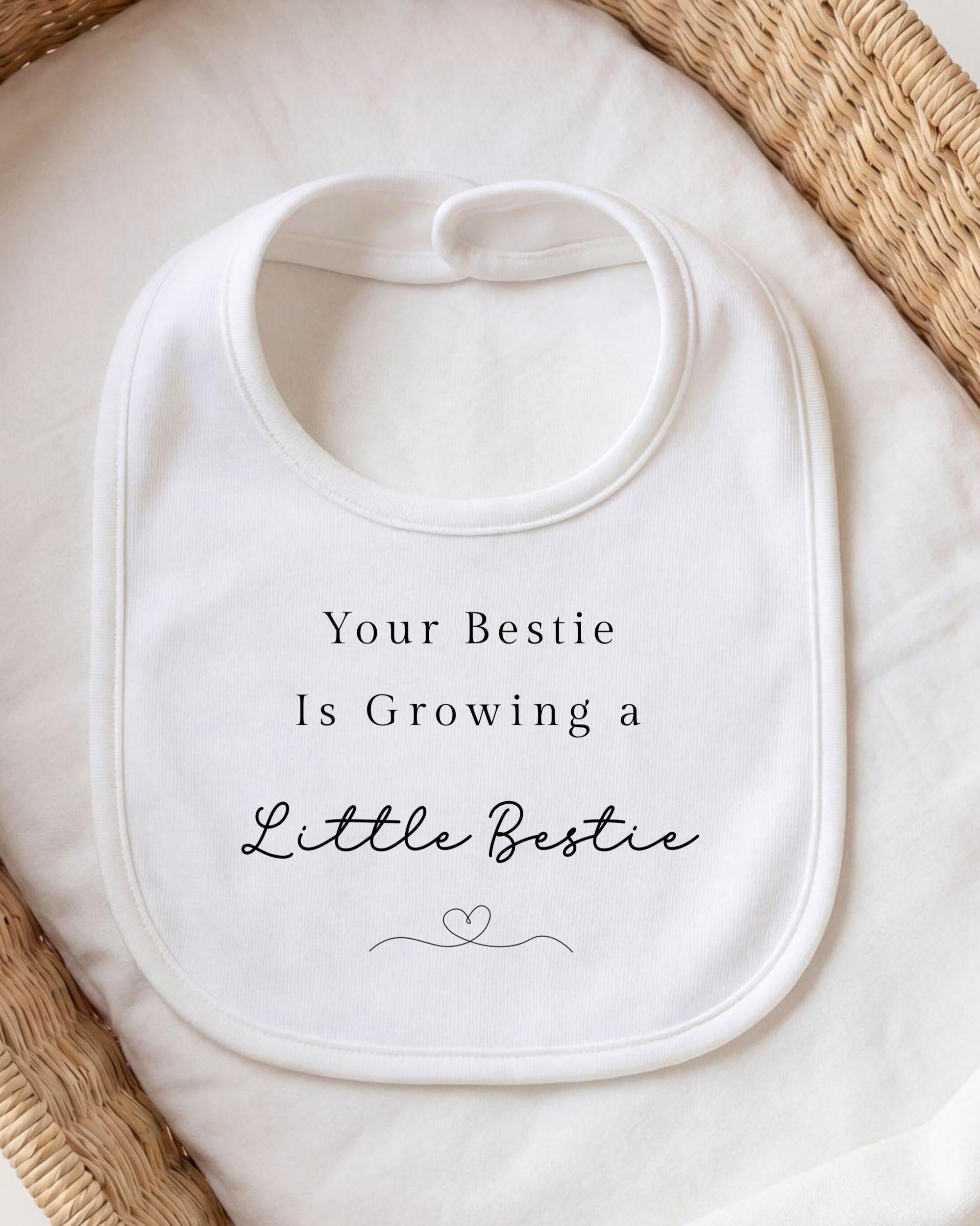 A white bib with the text "Your Bestie Is Growing a Little Bestie" printed on it, placed on a white cloth.
