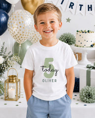 A young boy wearing a white t-shirt with the number 5 and the name "Oliver" printed on it, smiling at the camera. The background includes birthday decorations, balloons, and a cake.