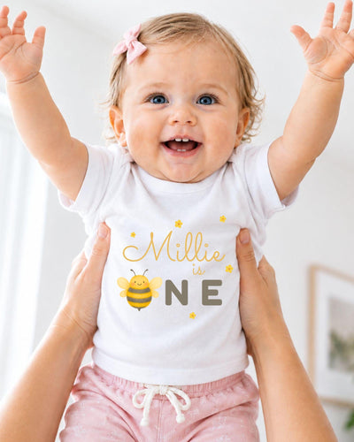 A smiling baby girl wearing a white t-shirt with a bee graphic and the text "Milly is one" is being held up by someone's arms.