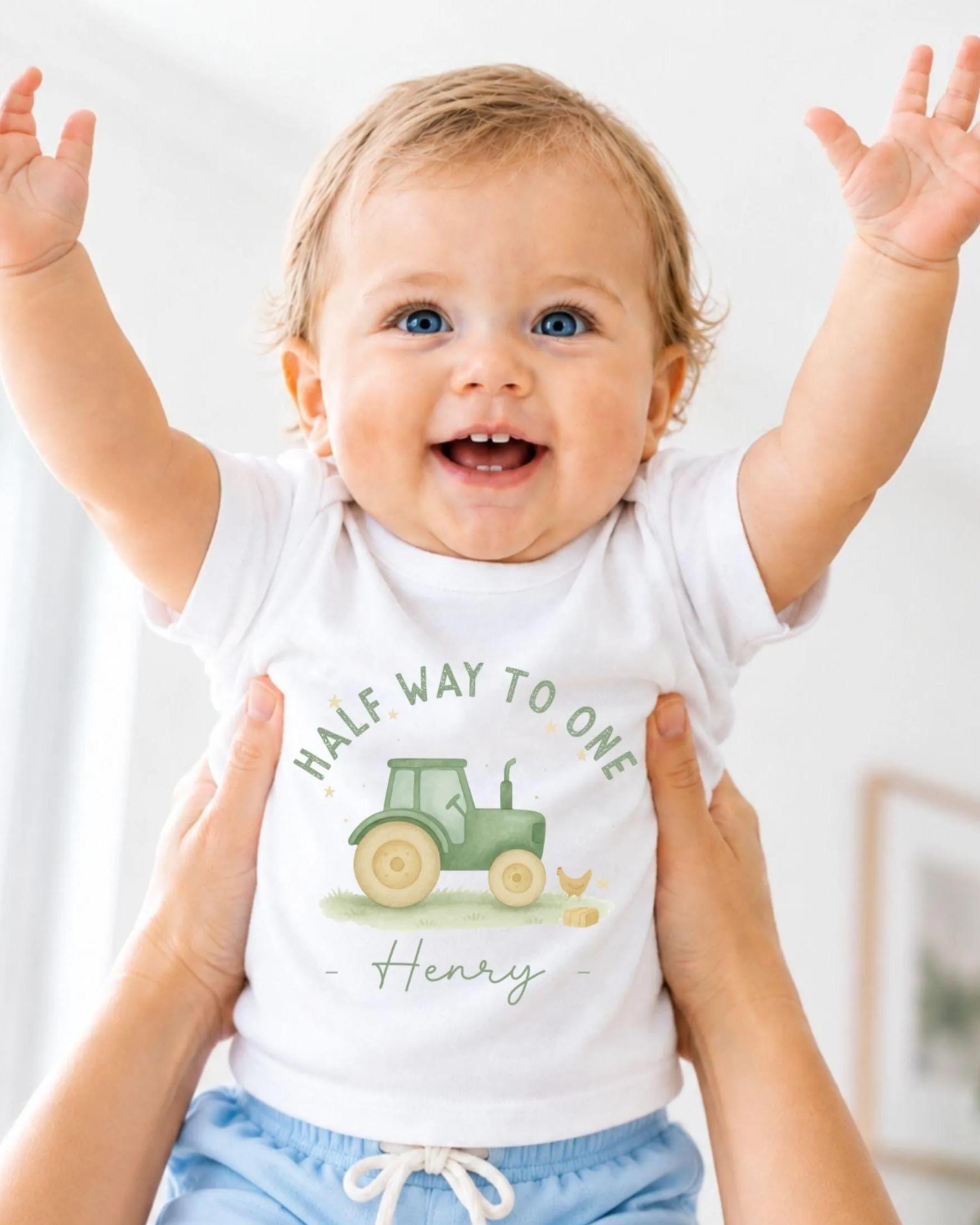 A smiling baby wearing a white t-shirt with a tractor graphic and the name "Henry" printed on it, being held up by someone's arms.