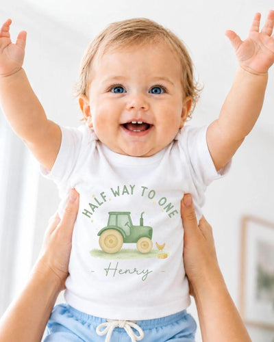 A smiling baby wearing a white t-shirt with a tractor graphic and the name "Henry" printed on it, being held up by someone's arms.