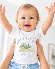 A smiling baby wearing a white t-shirt with a tractor graphic and the name "Henry" printed on it, being held up by someone's arms.