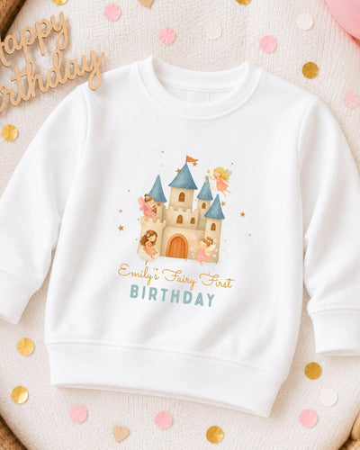 A white sweatshirt with a castle and fairy characters printed on it, along with the text "Emilly's Fairy First Birthday".