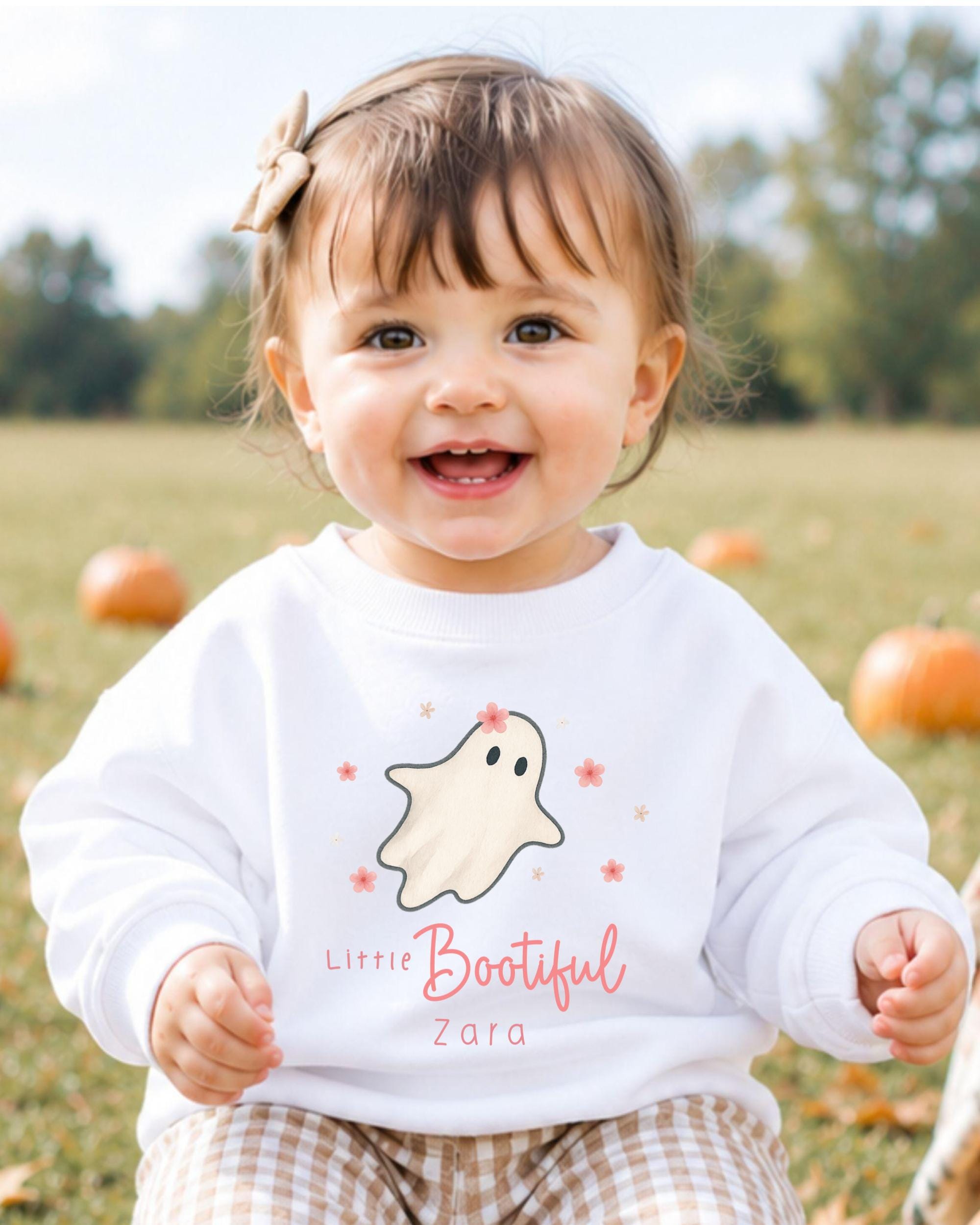 A smiling baby girl wearing a white sweater with a ghost and flower design, sitting in a field with pumpkins in the background.