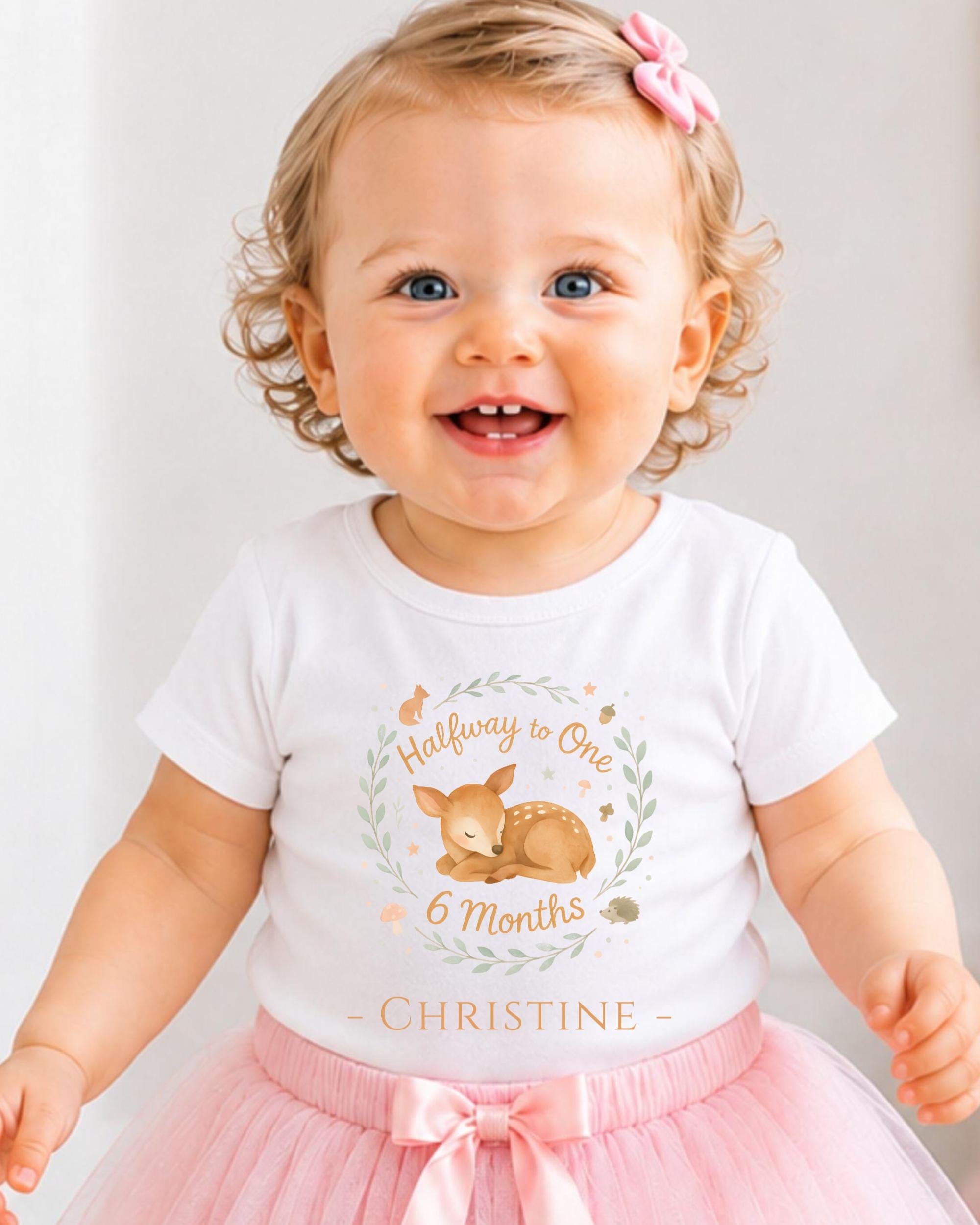 A smiling baby girl wearing a white t-shirt with a deer graphic and the text "Halfway to One 6 Months Christine" printed on it.