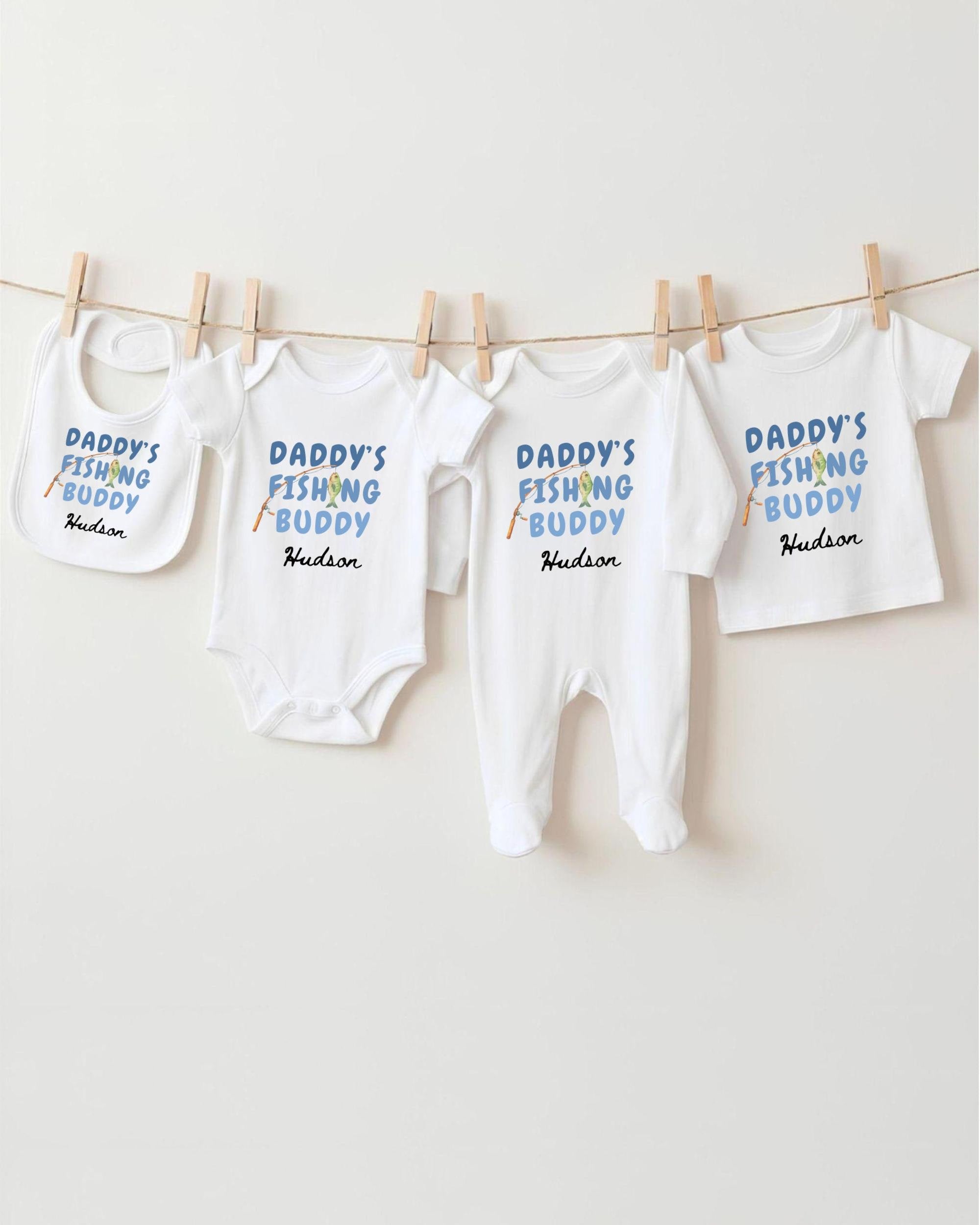 A row of white baby clothes with the text "DADMY'S FISHING BUDDY" printed on them, along with the name "Hudson" below.