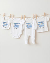 A row of white baby clothes with the text "DADMY'S FISHING BUDDY" printed on them, along with the name "Hudson" below.