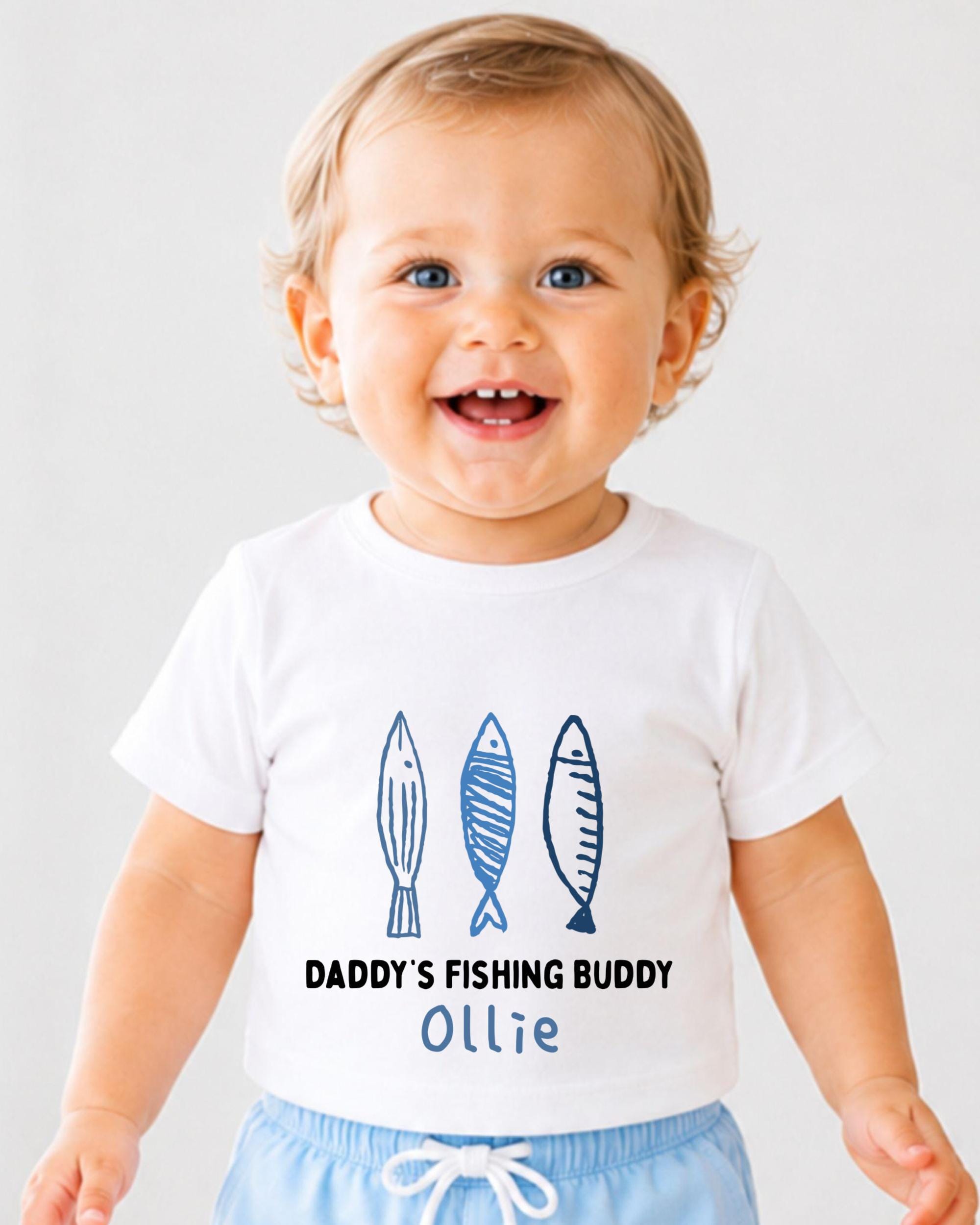 A smiling baby boy wearing a white t-shirt with a drawing of three fish and the text "DADDY'S FISHING BUDDY OLLIE" printed on it.
