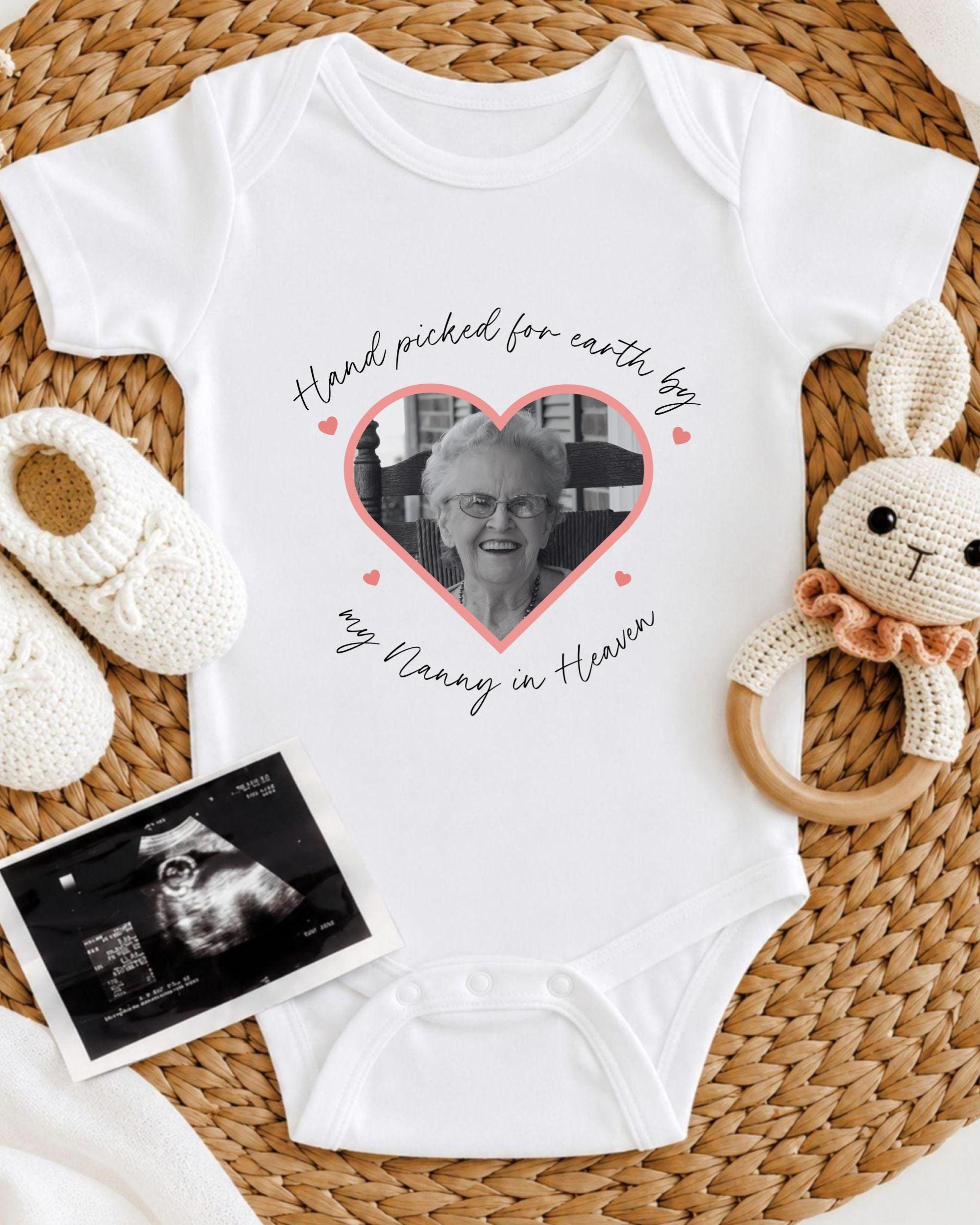 A white baby onesie with a heart-shaped photo of an elderly woman, surrounded by baby booties and a crocheted rabbit toy.