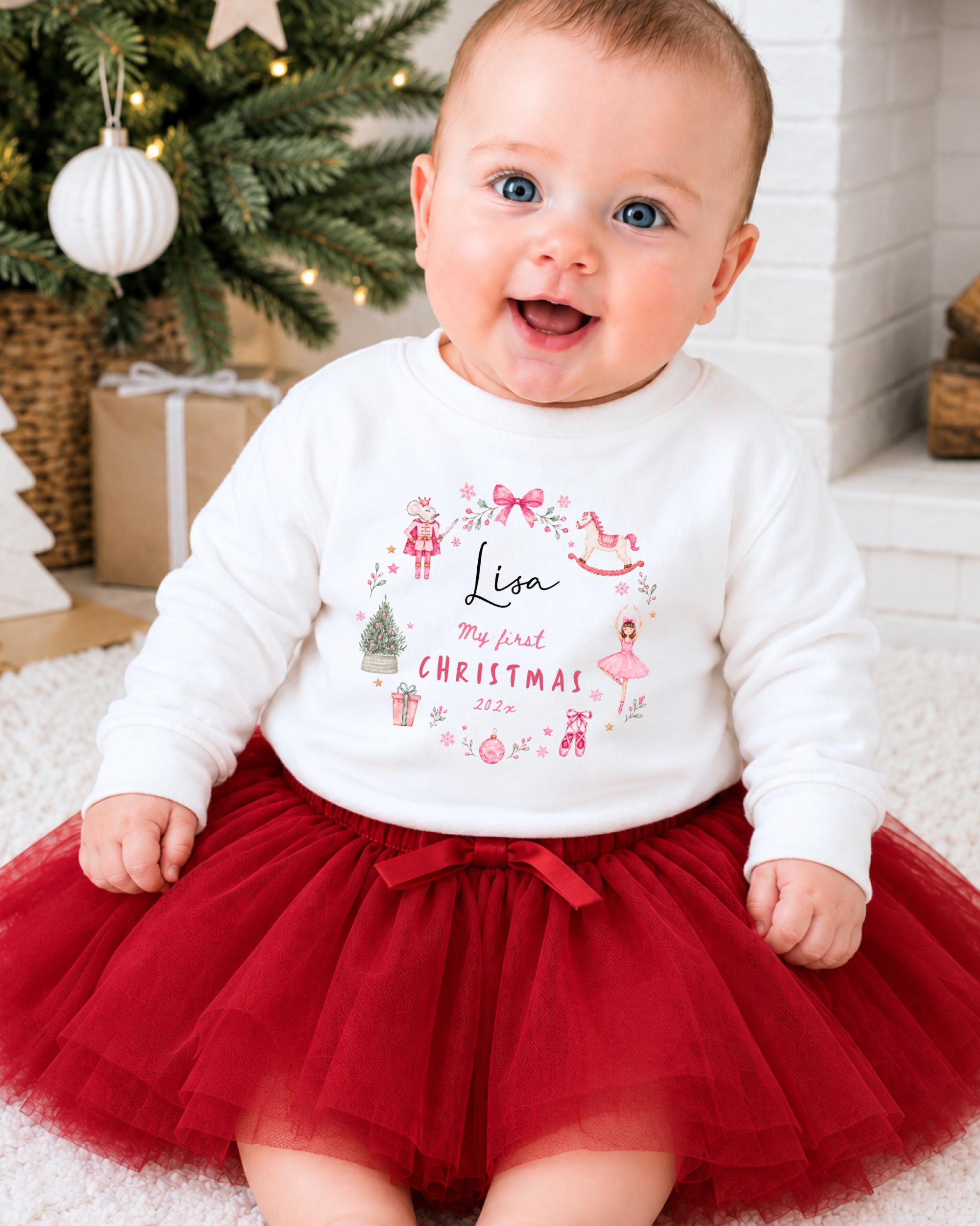 A smiling baby girl wearing a white sweater with a personalized design and a red tulle skirt, sitting in front of a Christmas tree.