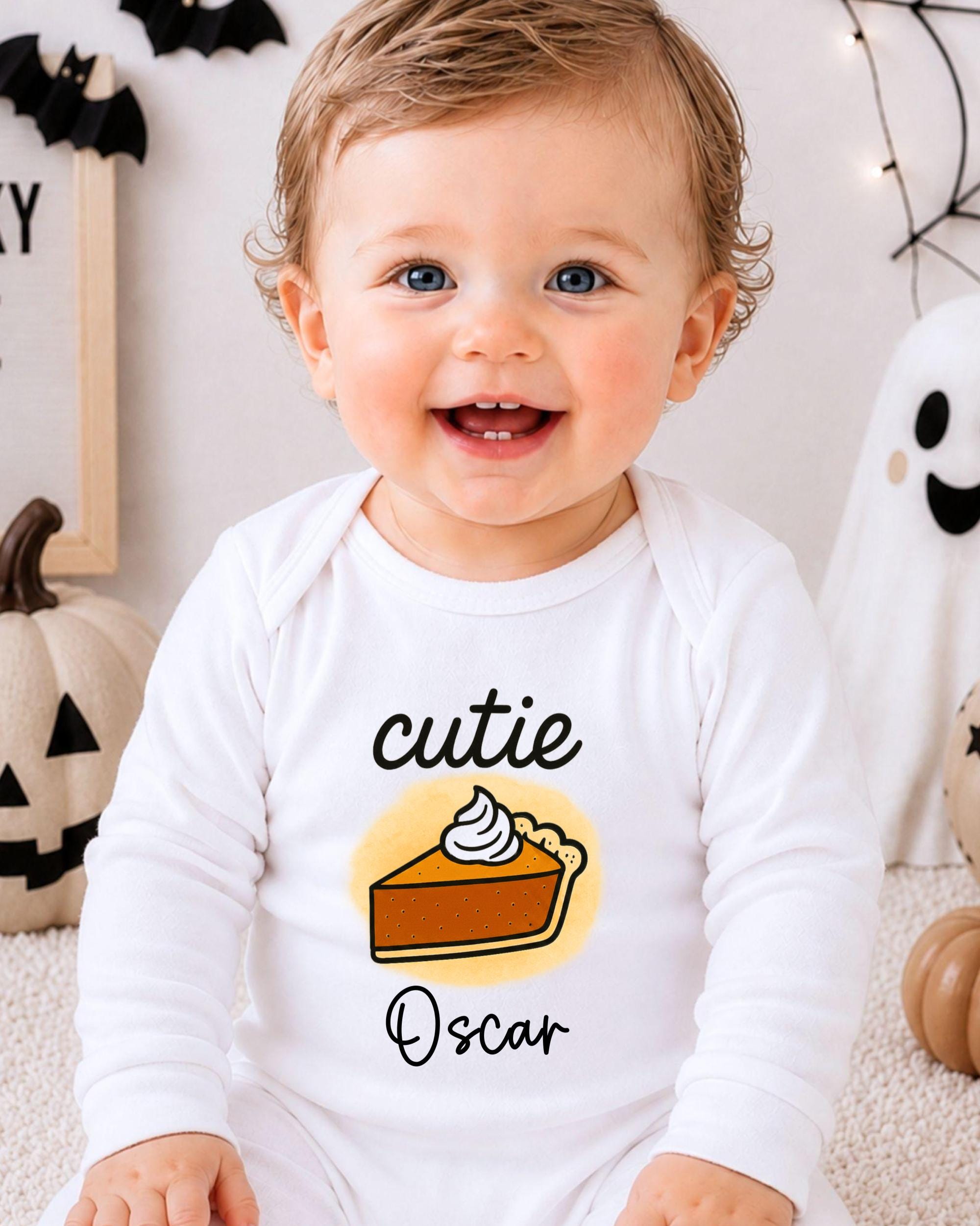 A smiling baby boy wearing a white onesie with a pumpkin and pie graphic, sitting in front of Halloween decorations.