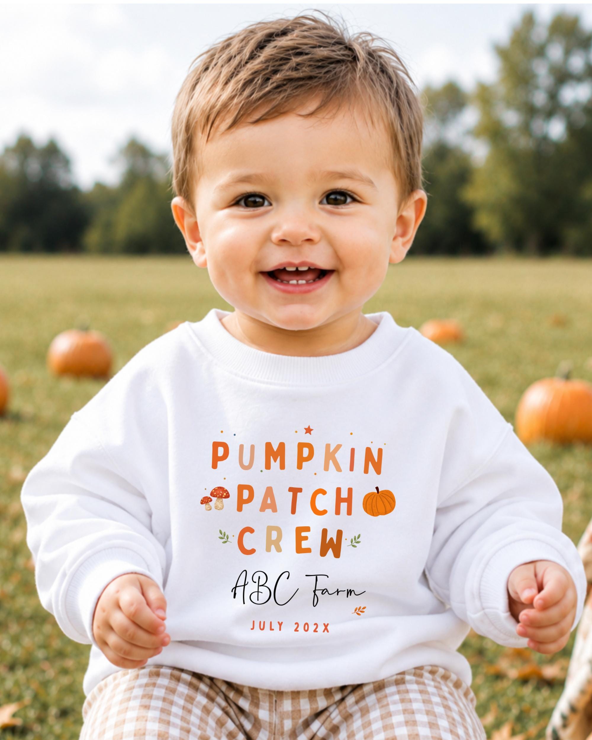 A smiling baby boy wearing a white sweatshirt with text and graphics related to a pumpkin patch crew.