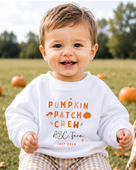 A smiling baby boy wearing a white sweatshirt with text and graphics related to a pumpkin patch crew.