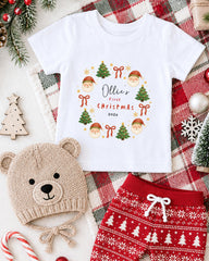 A white t-shirt with a Christmas-themed design, a beige knit hat with a Christmas tree and Santa Claus, and red and white patterned shorts, all arranged on a plaid blanket with Christmas decorations.