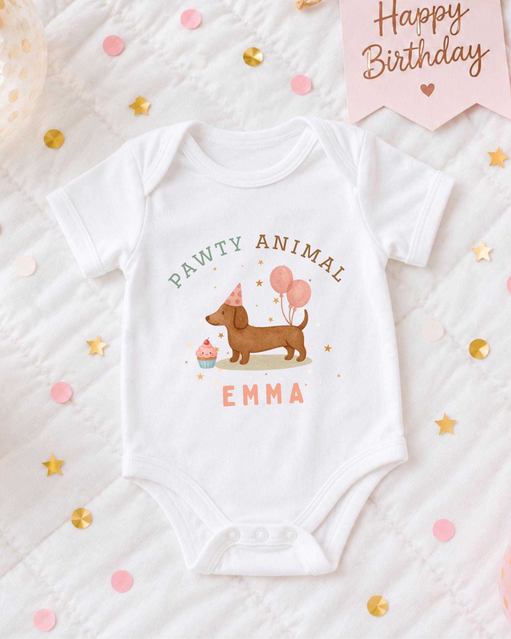 A white baby onesie with a dog and birthday party decorations, including a pink banner that says "Happy Birthday" and confetti.