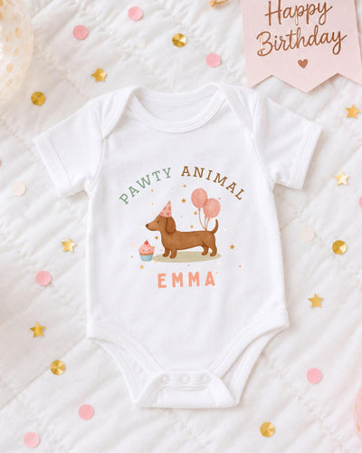 A white baby bodysuit with a dog and birthday party decorations, including a pink banner that says "Happy Birthday" and confetti.