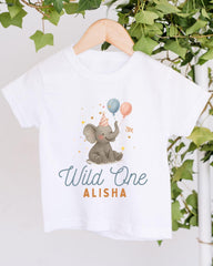 A white t-shirt with a graphic design of an elephant holding balloons and wearing a party hat, along with the text "Wild One Alisha" printed on it.