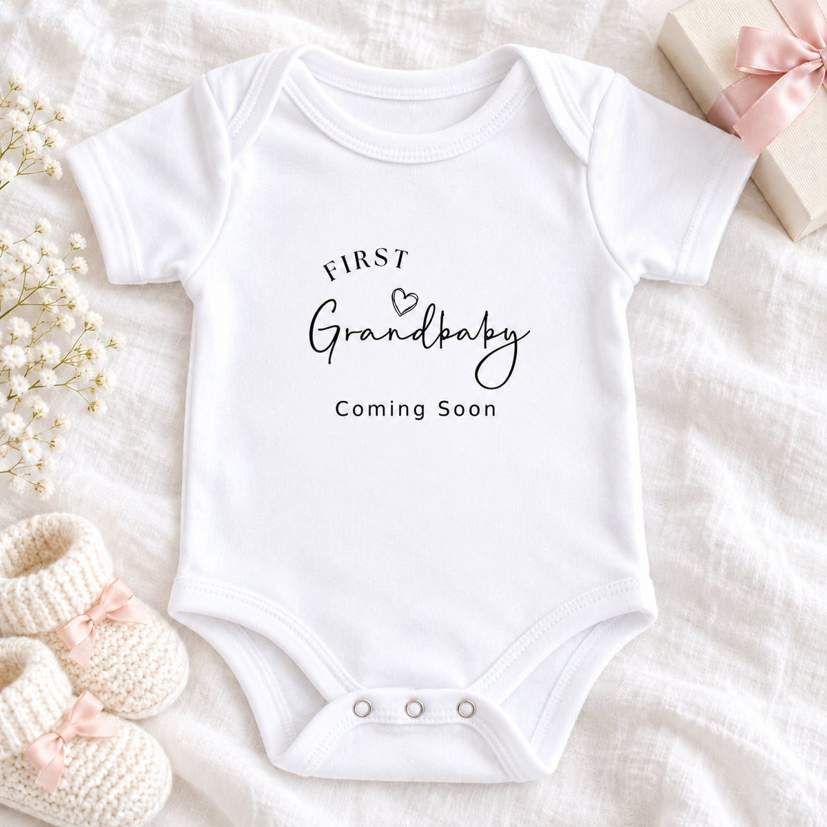 White First Grandbaby Coming Soon pregnancy announcement Baby Vest with black script lettering, styled with a cream gift box, ribbon, knitted baby booties and eucalyptus on beige linen, with props notice.