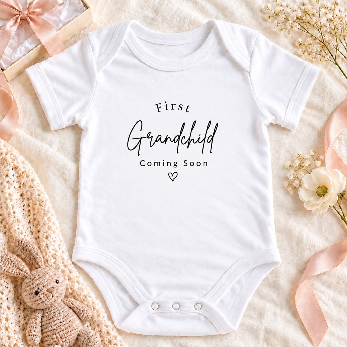 Square pregnancy announcement photo of a white First Grandchild Coming Soon Baby Vest with cream gift box, ribbon, baby booties and eucalyptus on beige linen.