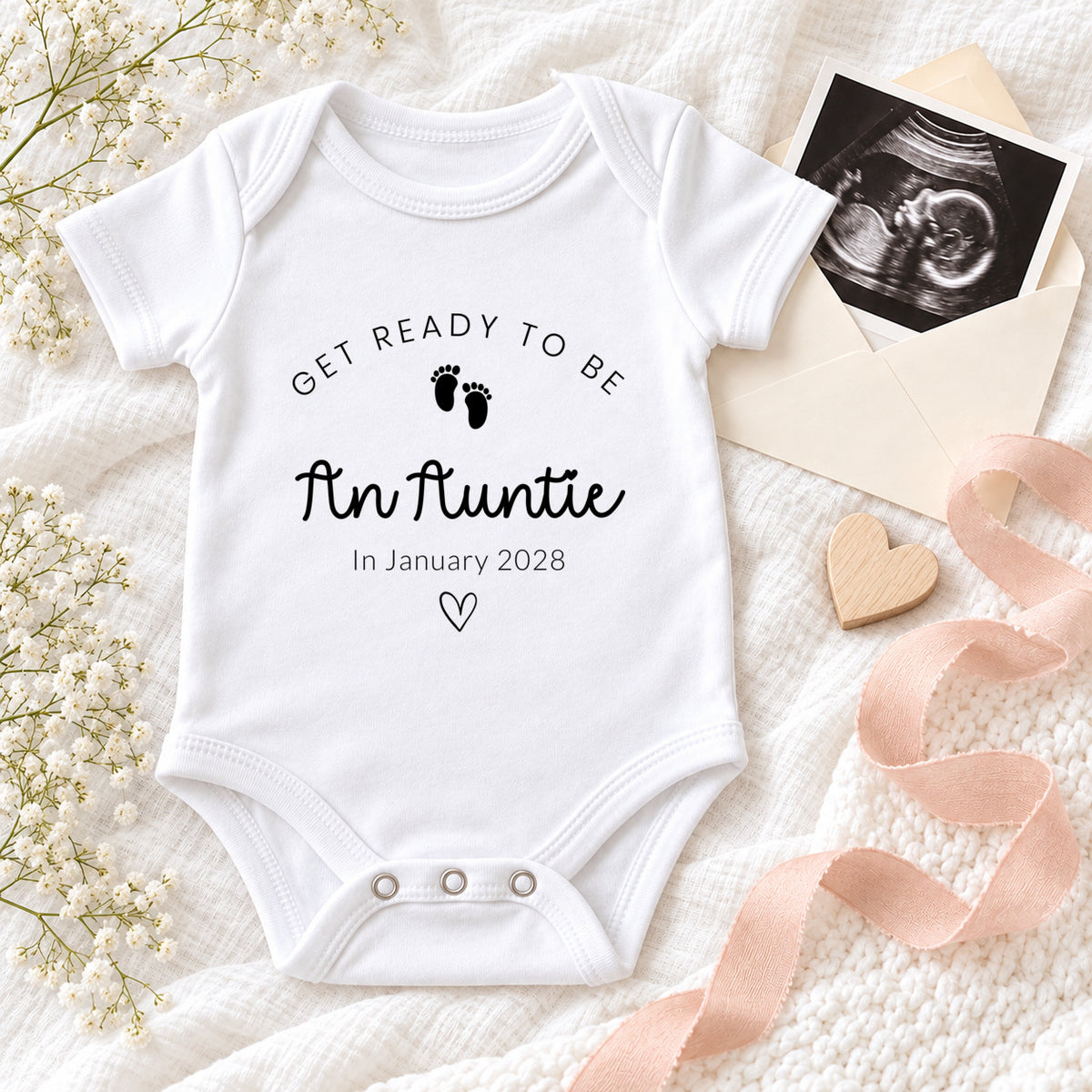 White Baby Vest printed with Get Ready to Be An Auntie, tiny footprints, In January 2028 and a heart, styled with a scan photo, cream envelope, blush ribbon and flowers.
