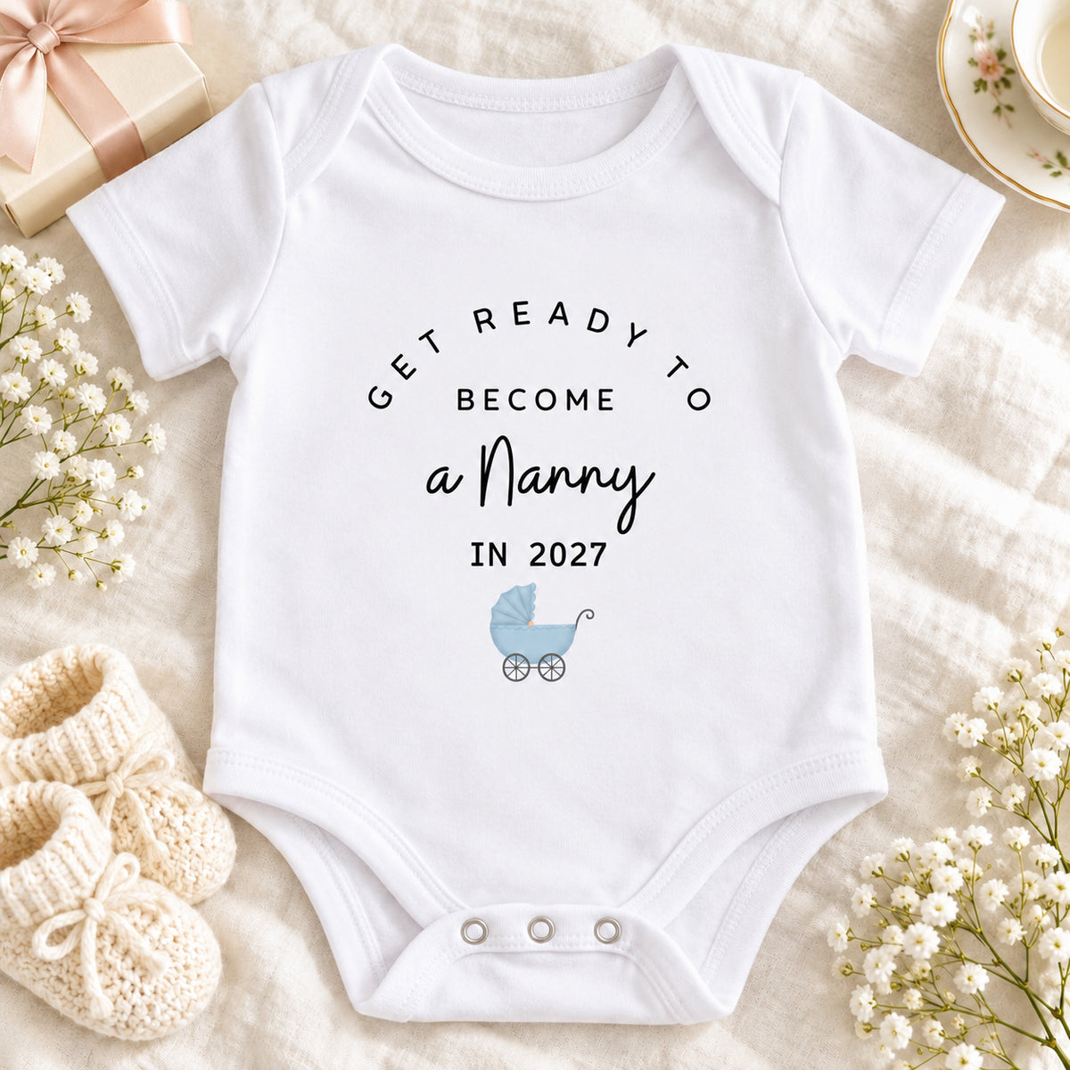 Square pregnancy announcement image of a white Get Ready to Become a Nanny Baby Vest with pale blue pram, styled on beige linen with a gift box, ribbon, baby booties, teacup and dried flowers.