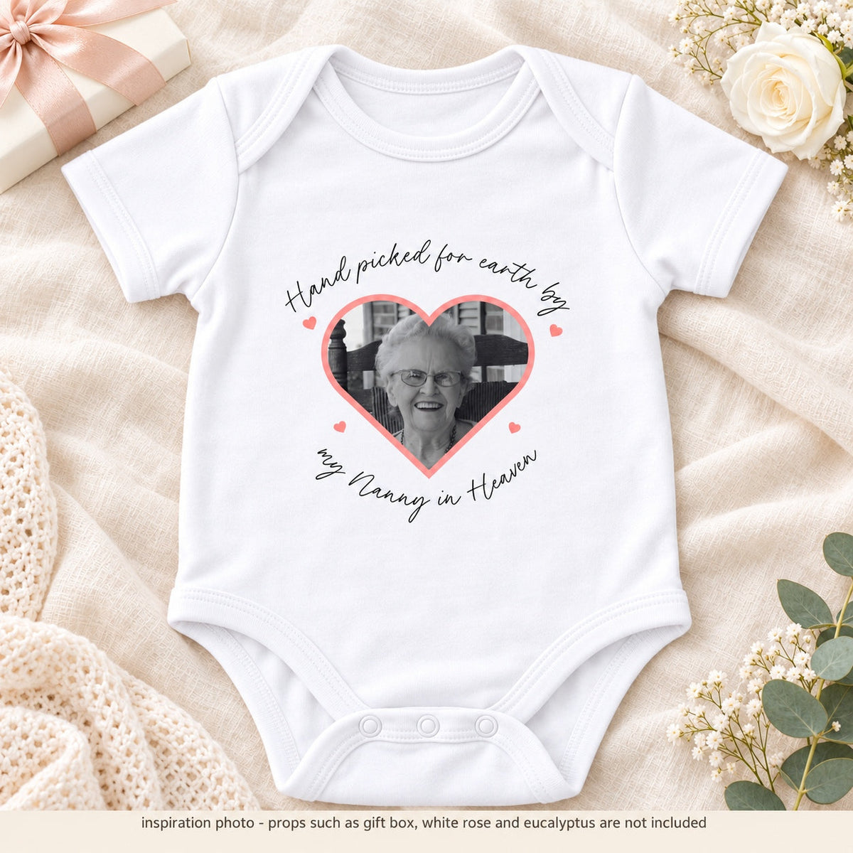 Personalised white memorial pregnancy announcement Baby Vest with a heart-shaped black and white photograph, remembrance wording, cream gift box, white rose and eucalyptus on beige linen, with props notice.