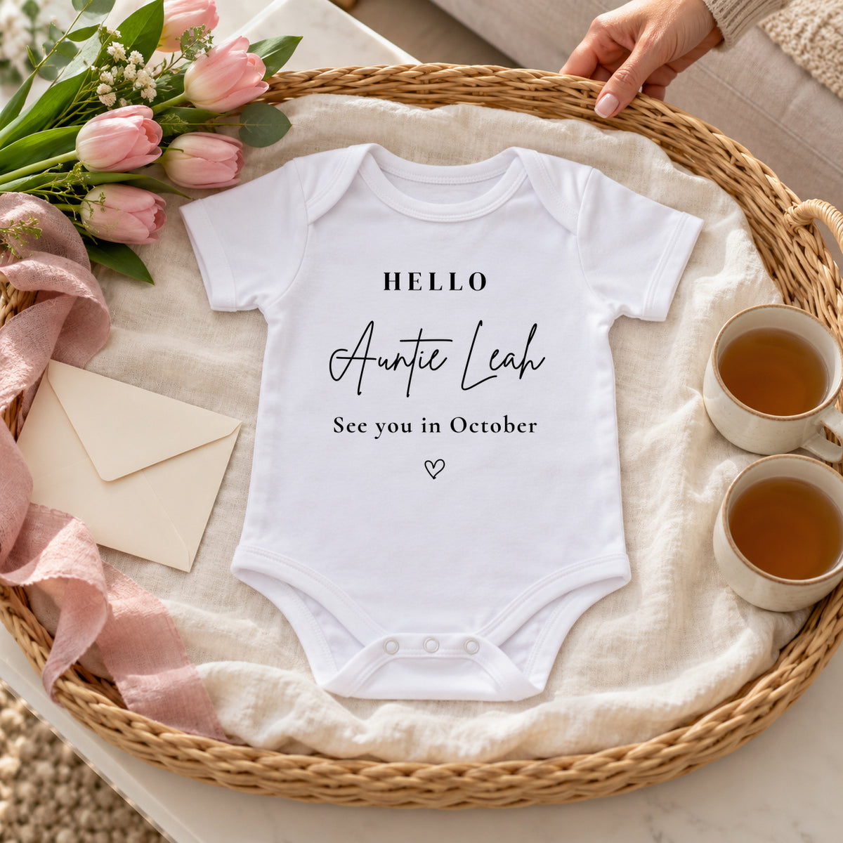 White Hello Auntie Leah See you in October Baby Vest on an ivory-lined wicker tray with two cups of tea, blush tulips, ribbon and a cream envelope.