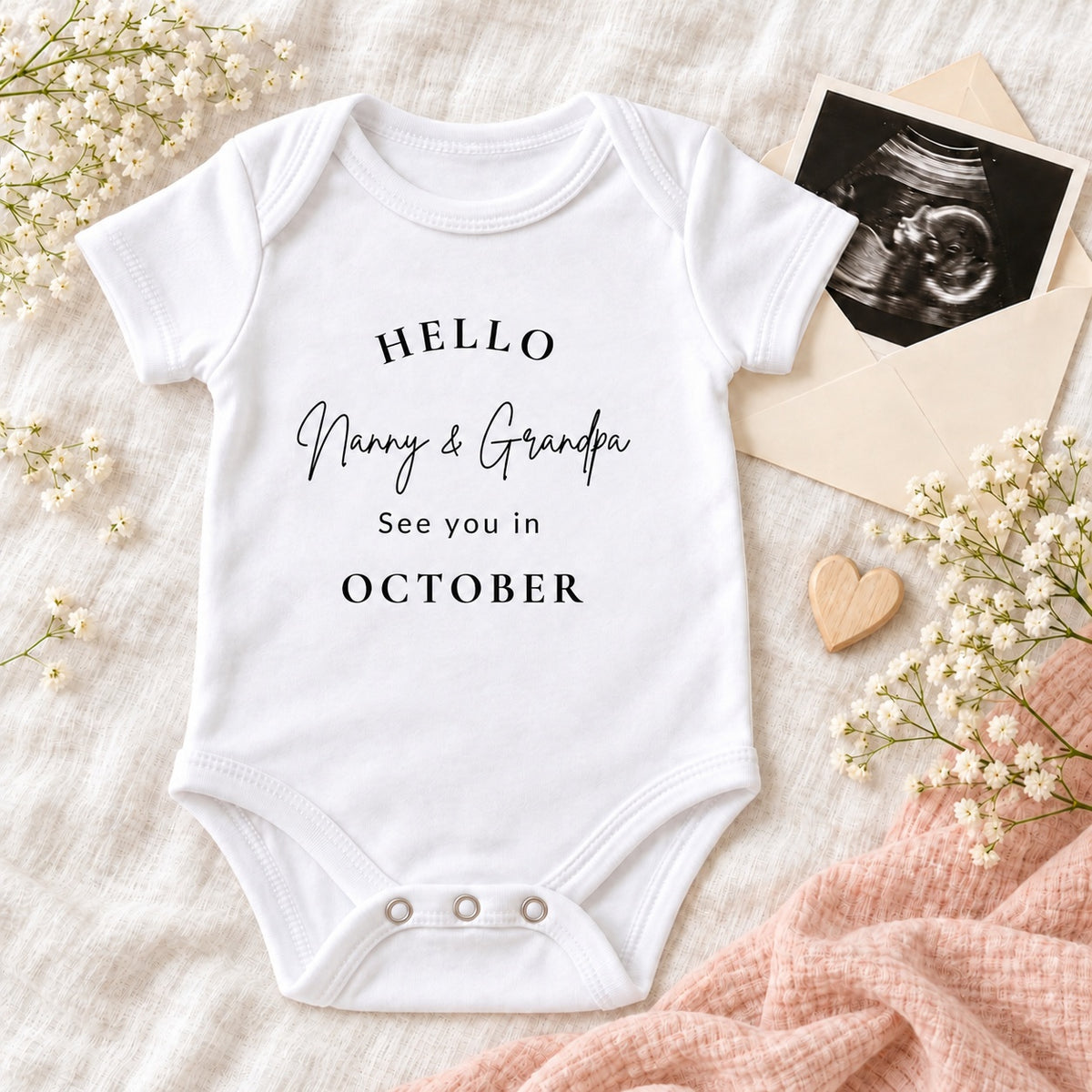 White Baby Vest printed with Hello Nanny & Grandpa, See you in October, styled with a scan photo, ivory envelope, wooden heart, blush muslin and delicate white flowers.