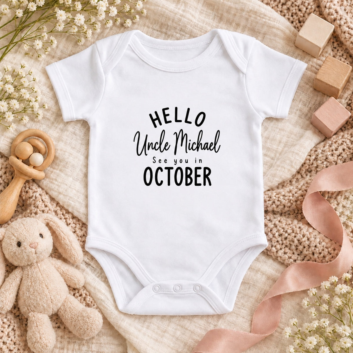 White Baby Vest printed with HELLO Uncle Michael See you in OCTOBER, styled with ivory knit, blush ribbon, white flowers and wooden nursery toys