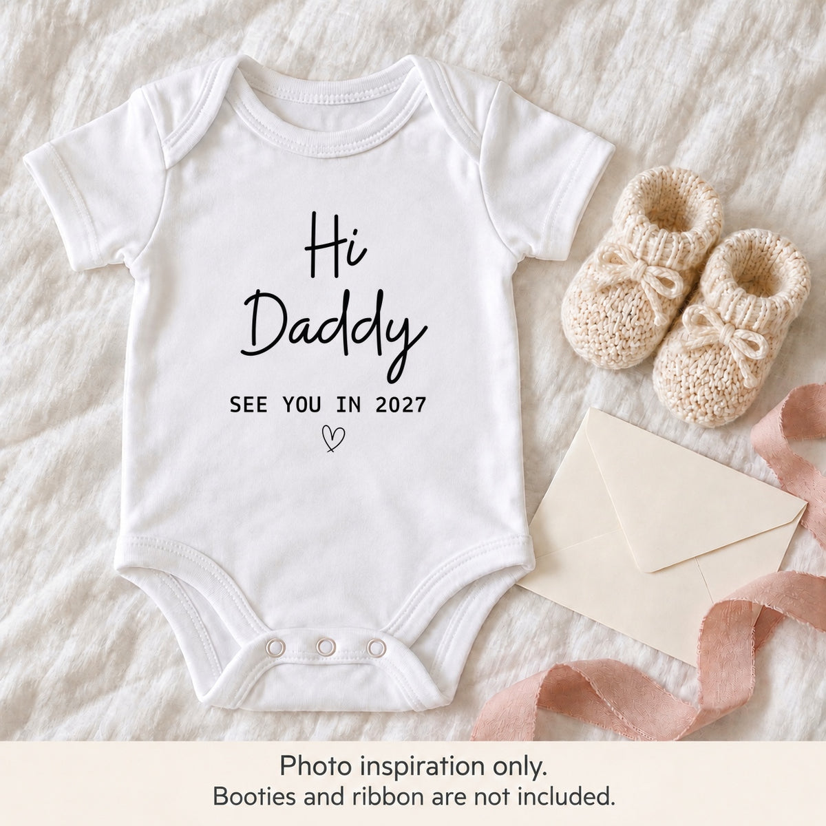White Hi Daddy pregnancy announcement baby vest on ivory linen beside knitted baby booties, a blank envelope and blush pink ribbon.