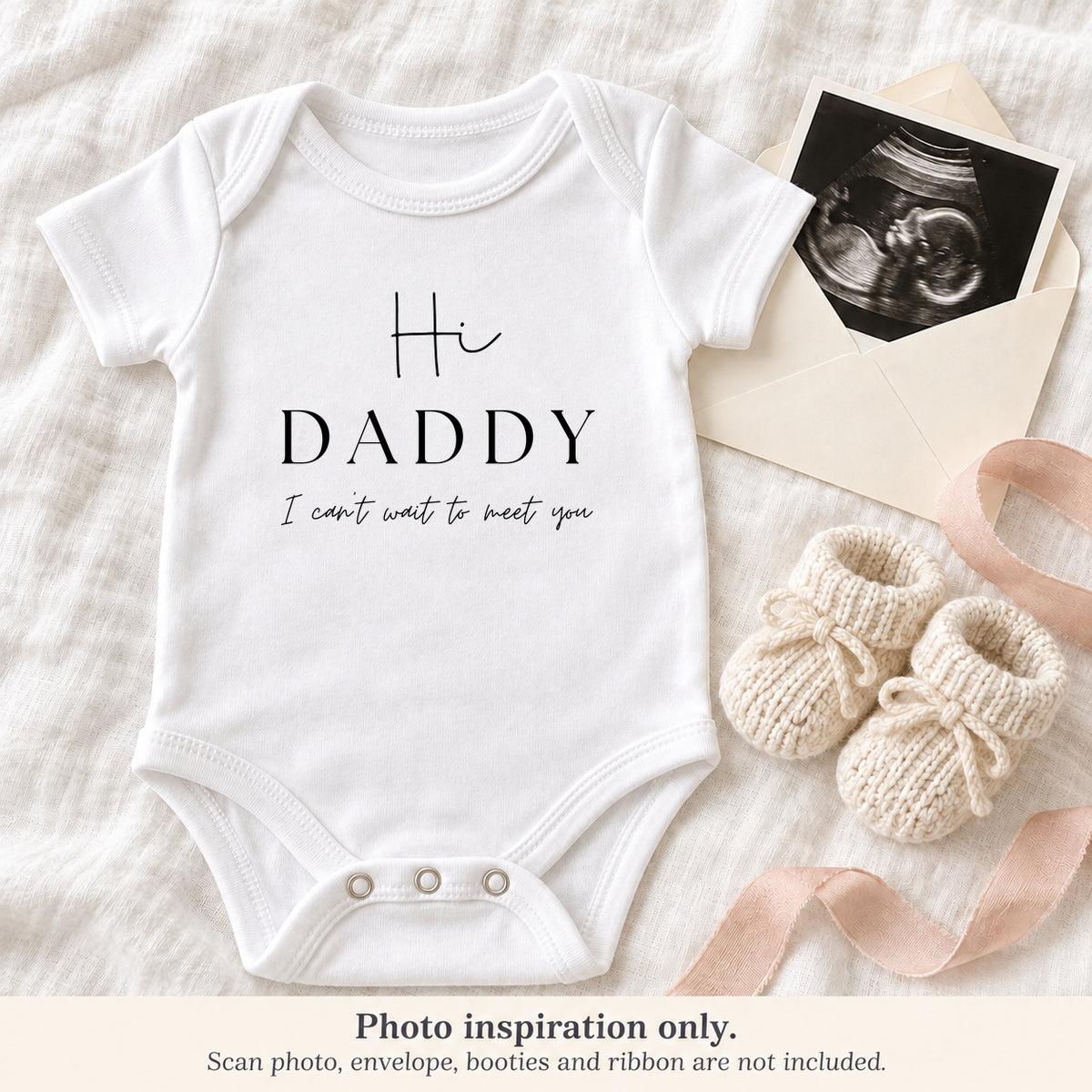 White 'Hi Daddy I Can't Wait to Meet You' pregnancy announcement baby vest beside an ultrasound photo, ivory envelope, cream booties and blush ribbon.