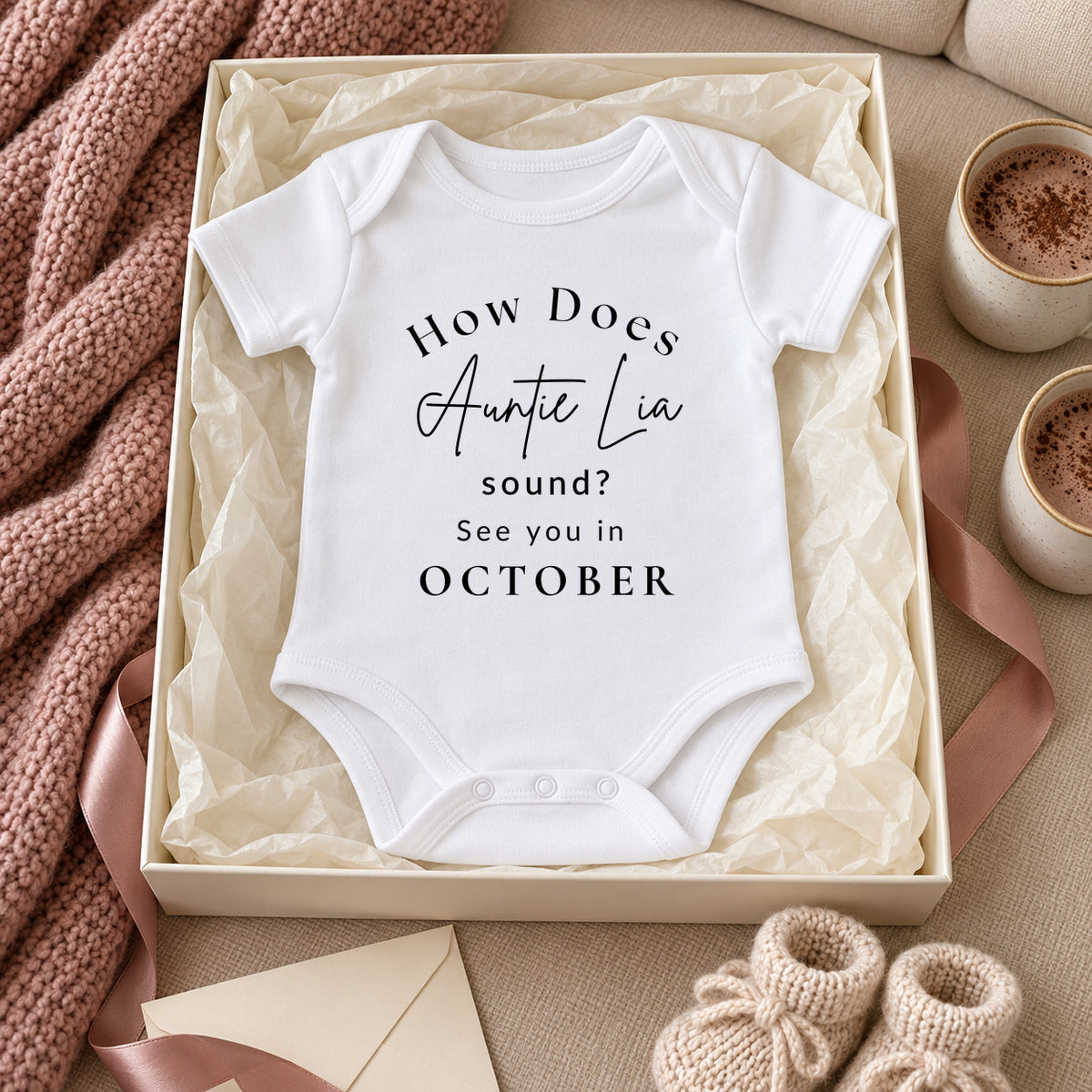 White How Does Auntie Lia sound See you in OCTOBER Baby Vest in an ivory gift box beside hot chocolate, a rose throw, ribbon and baby booties.