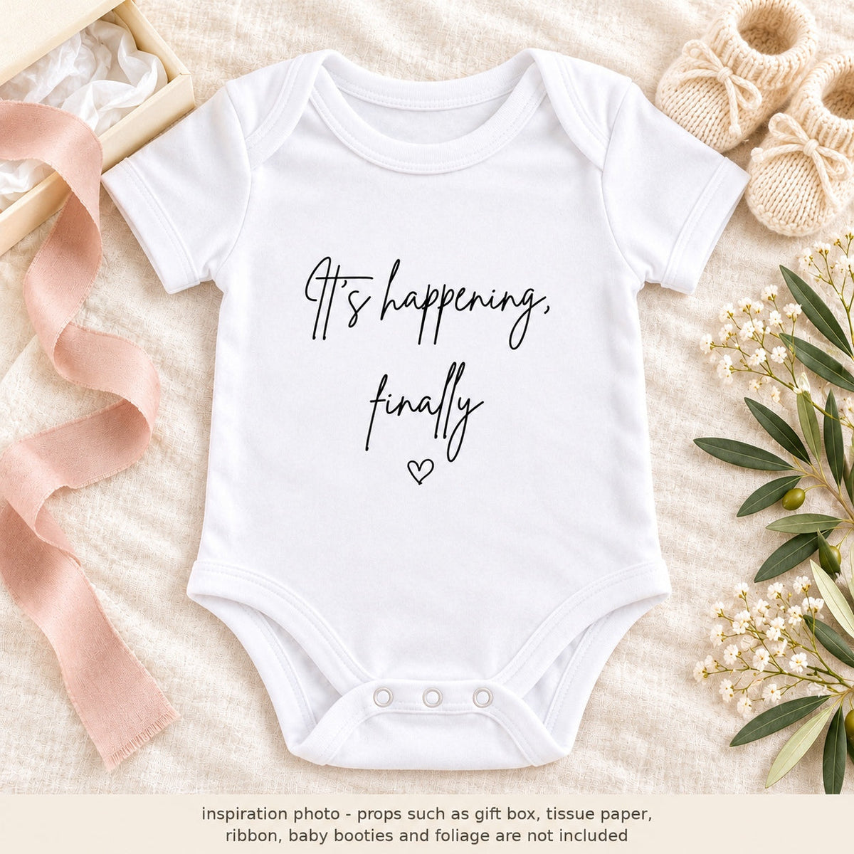 Pregnancy announcement flat lay with a white It's happening, finally Baby Vest, cream gift box, tissue paper, blush ribbon, knitted baby booties and olive foliage, with a props notice.
