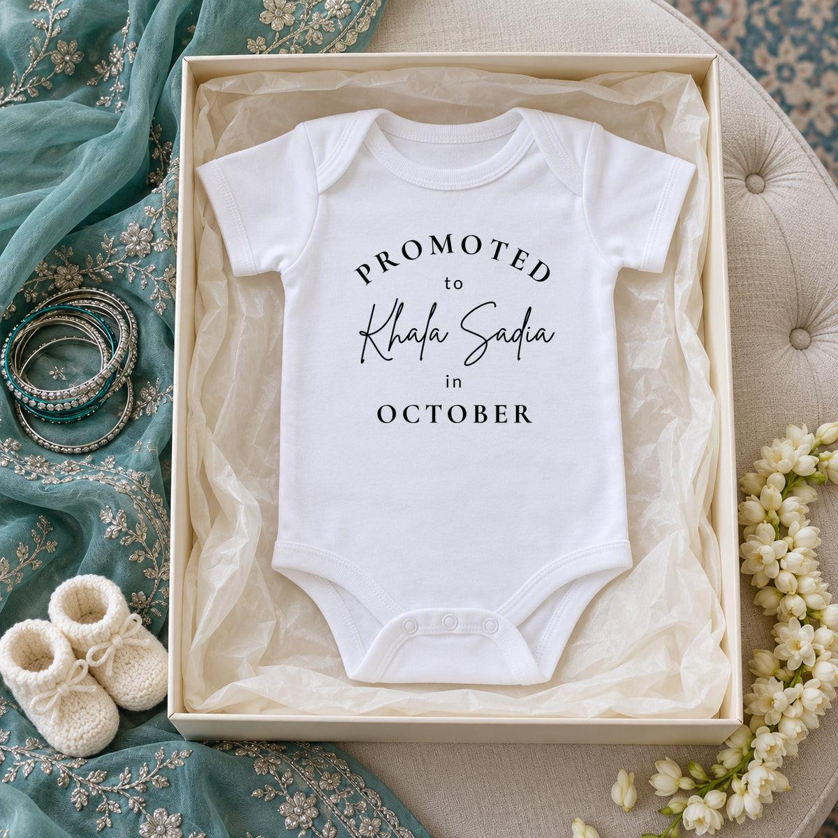 White Baby Vest reading Promoted to Khala Sadia in October inside an ivory gift box on a cream ottoman, with teal embroidered fabric, silver and teal bangles, jasmine and knitted baby booties.