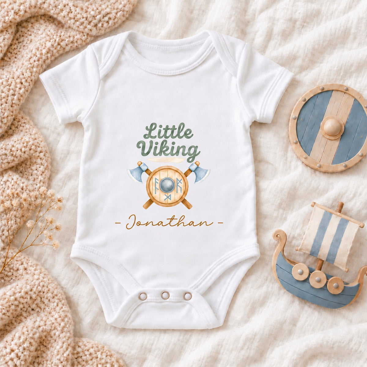 Square flat lay of a white personalised Little Viking Baby Vest with shield and axes artwork, Jonathan name, wooden Viking shield and toy longship.