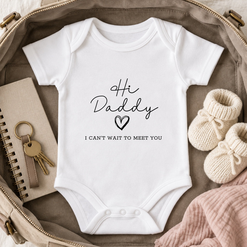 White Hi Daddy pregnancy announcement baby vest with a small black chest print in an open taupe work bag beside cream booties, a notebook and keys.