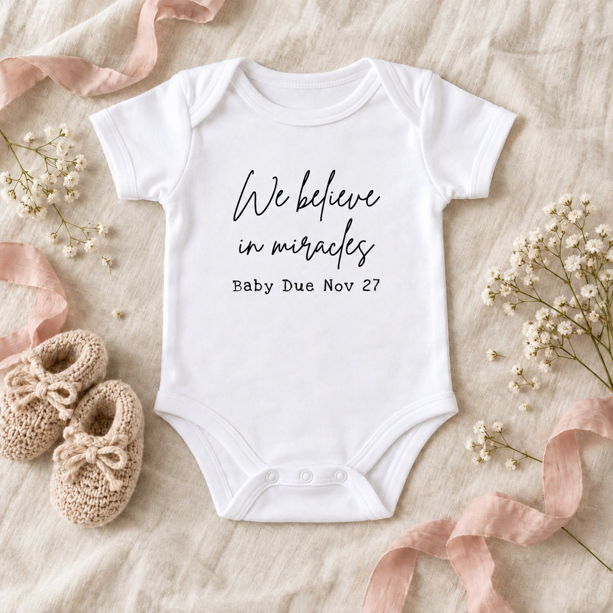 White Baby Vest with black 'We believe in miracles' and 'Baby Due Nov 27' artwork on ivory linen beside beige knitted booties, blush ribbon and white flowers.