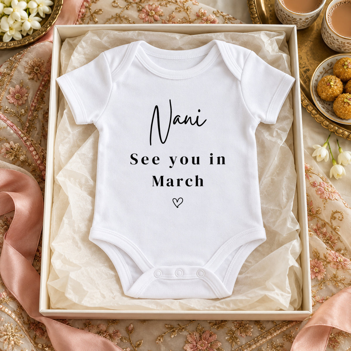 White Nani Baby Vest reading 'Nani See you in March' in an ivory gift box on an embroidered dupatta, beside chai, ladoo sweets and jasmine.