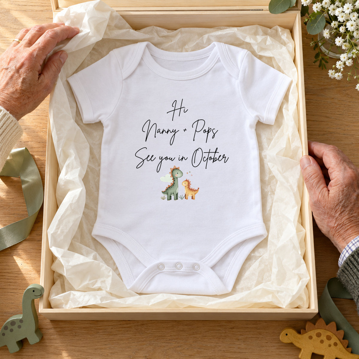 Grandparents opening a wooden keepsake box with a white dinosaur Baby Vest reading Hi Nanny + Pops, See you in October, with sage ribbon and wooden dinosaurs.