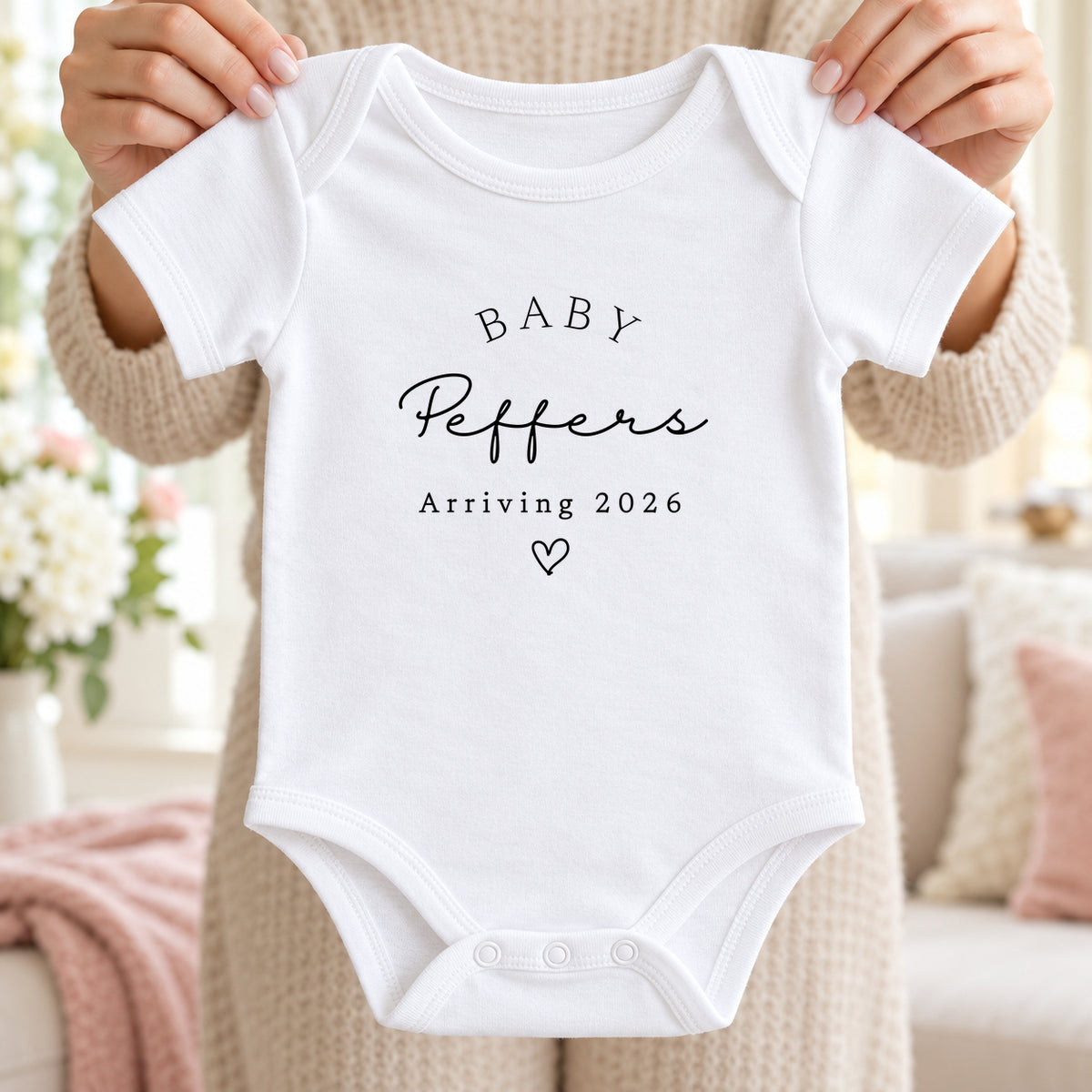 Adult hands holding a white Baby Vest reading Baby Peffers, Arriving 2026, with a heart, in an airy cream living room with blush details.
