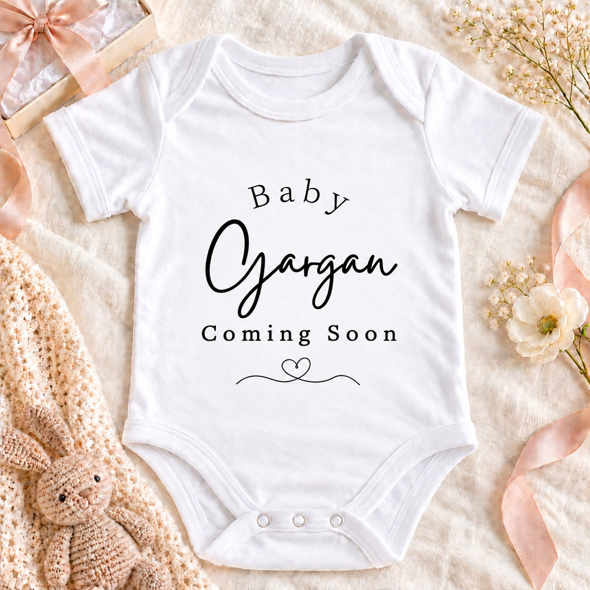 Square pregnancy announcement image of a white Baby Gargan Coming Soon Baby Vest with heart flourish, cream envelope, beige ribbon and ultrasound photograph on neutral linen.