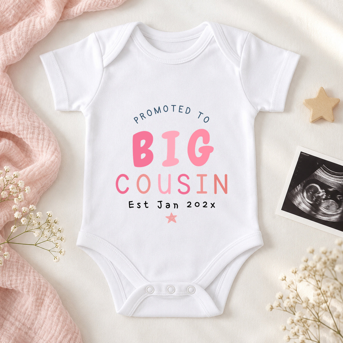 White Promoted to Big Cousin Baby Vest with pink lettering and Est Jan 202x, styled with blush muslin, a wooden star, white flowers and an ultrasound scan.
