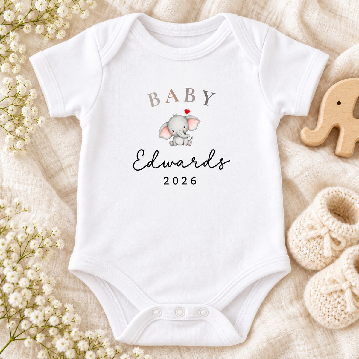 White Baby Vest with BABY Edwards 2026 text, a grey elephant and red heart on ivory linen, beside a wooden elephant, cream knit and white flowers.