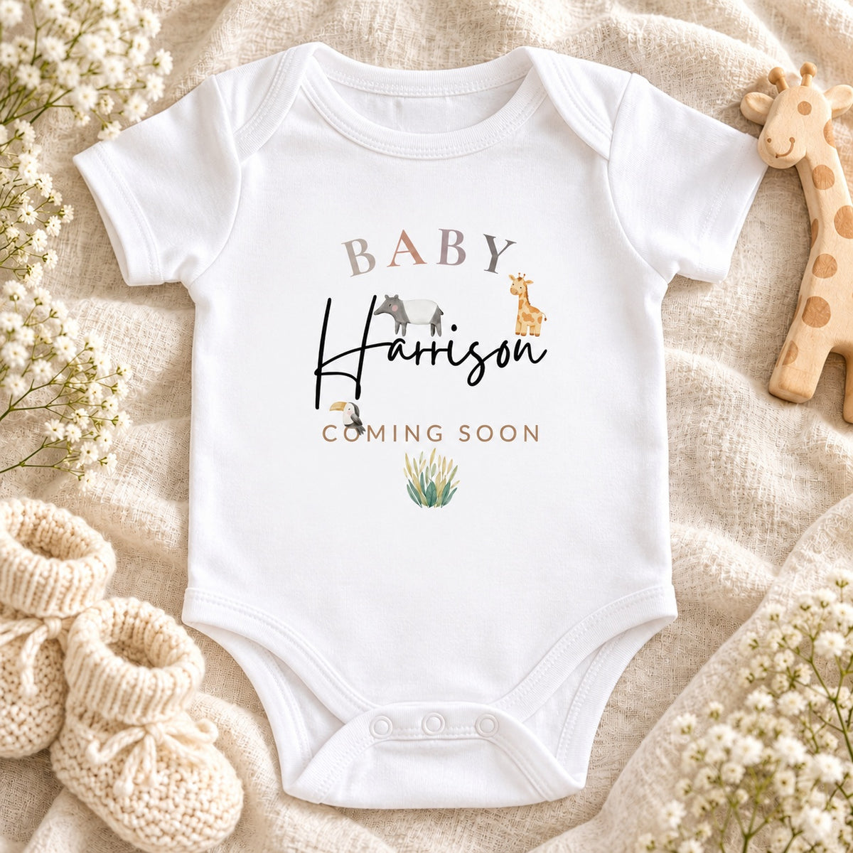 White Baby Vest with BABY Harrison Coming Soon safari artwork, three animals and foliage on ivory linen, with a wooden giraffe, cream booties and white flowers.