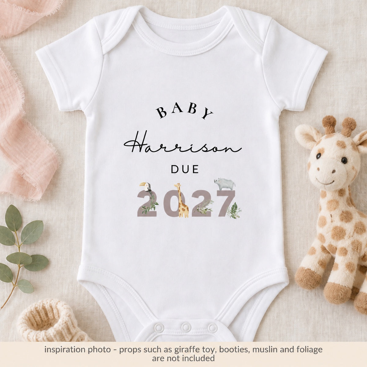 White safari pregnancy announcement baby vest reading Baby Harrison Due 2027 on ivory linen beside a plush giraffe, cream booties and blush fabric.